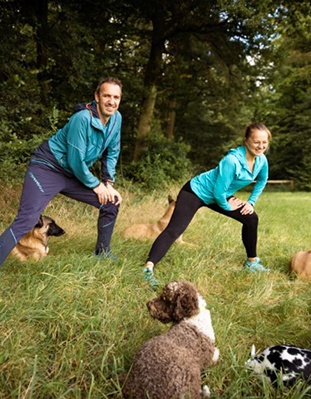 Nadine steht mit Jochen auf einer Wiese. Zusammen machen sie Dehnübungen und um sie herum liegen entspannt Hunde. Bild von Carolin Jütz