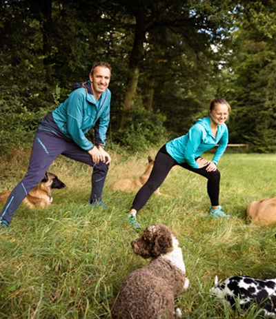 Nadine steht mit Jochen auf einer Wiese. Zusammen machen sie Dehnübungen und um sie herum liegen entspannt Hunde. Bild von Carolin Jütz