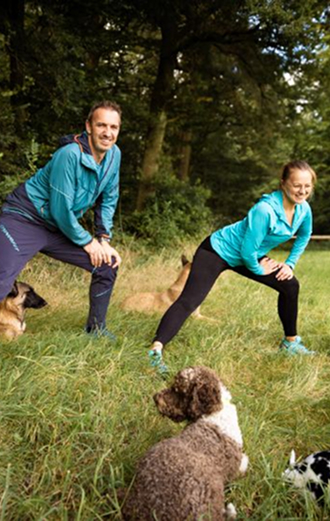 Nadine steht mit Jochen auf einer Wiese. Zusammen machen sie Dehnübungen und um sie herum liegen entspannt Hunde. Bild von Carolin Jütz